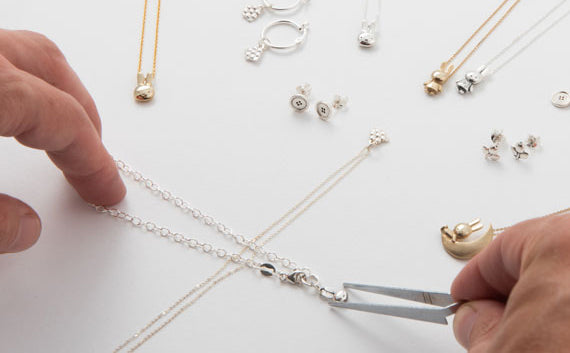Hands assembling a silver necklace with jewellery tools, surrounded by handmade gold and silver pieces