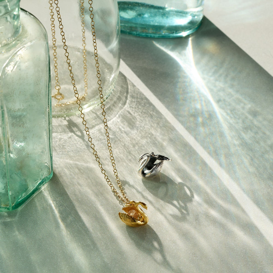 Blackbird Jewellery gold vermeil swan necklace and sterling silver swan charm displayed beside glass bottles in sunlight