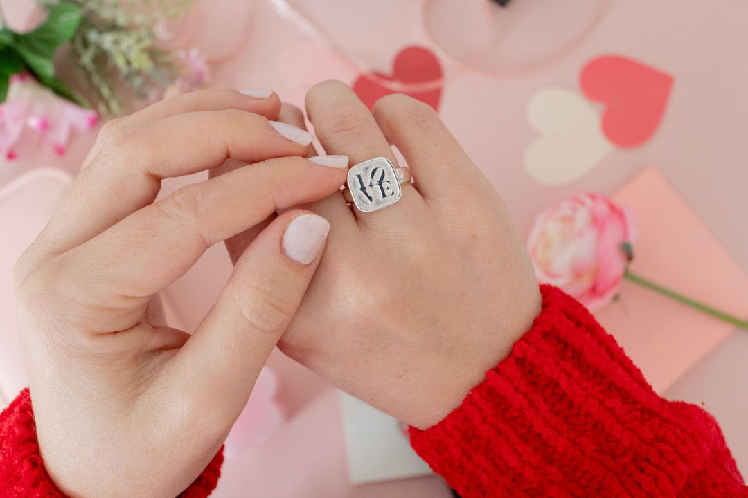 Hands wearing a sterling silver love ring against a soft pink Valentine’s Day background with heart decorations