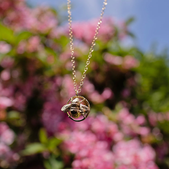 18ct gold vermeil queen bee necklace with detailed wings, front view with beautiful flowers in the background.