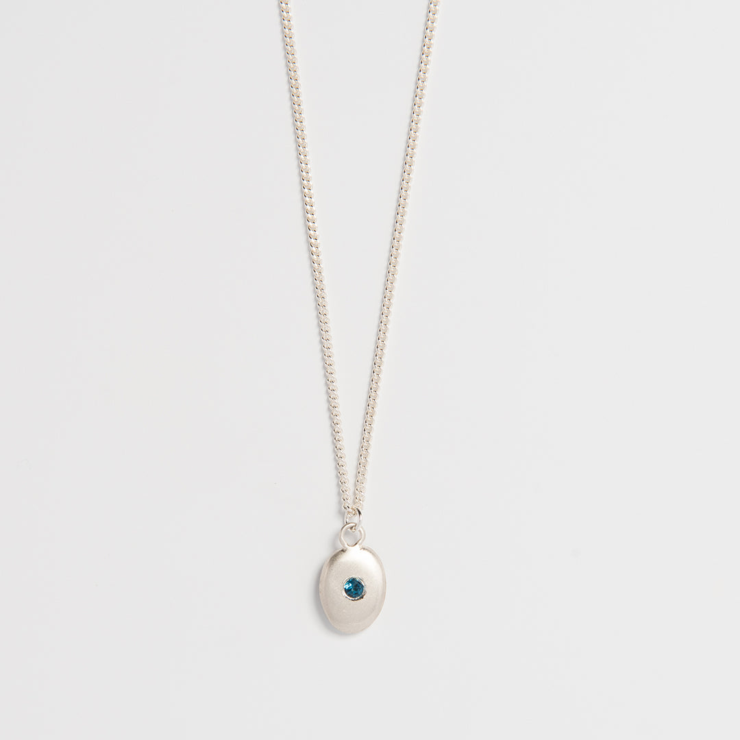 A sterling silver necklace with a small pebble-shaped pendant featuring a blue topaz at its center, suspended from a delicate sateen-coated chain.