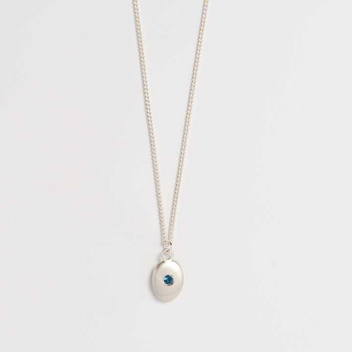 A sterling silver necklace with a small pebble-shaped pendant featuring a blue topaz at its center, suspended from a delicate sateen-coated chain.