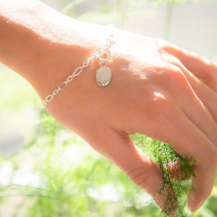 Blackbird Jewellery sterling silver bracelet with a pebble charm, being worn on a hand against a backdrop of greenery.