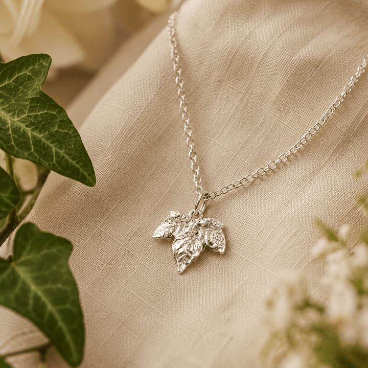 A silver ivy leaf pendant necklace resting on a textured beige fabric surface, styled with green ivy leaves and soft white flowers.