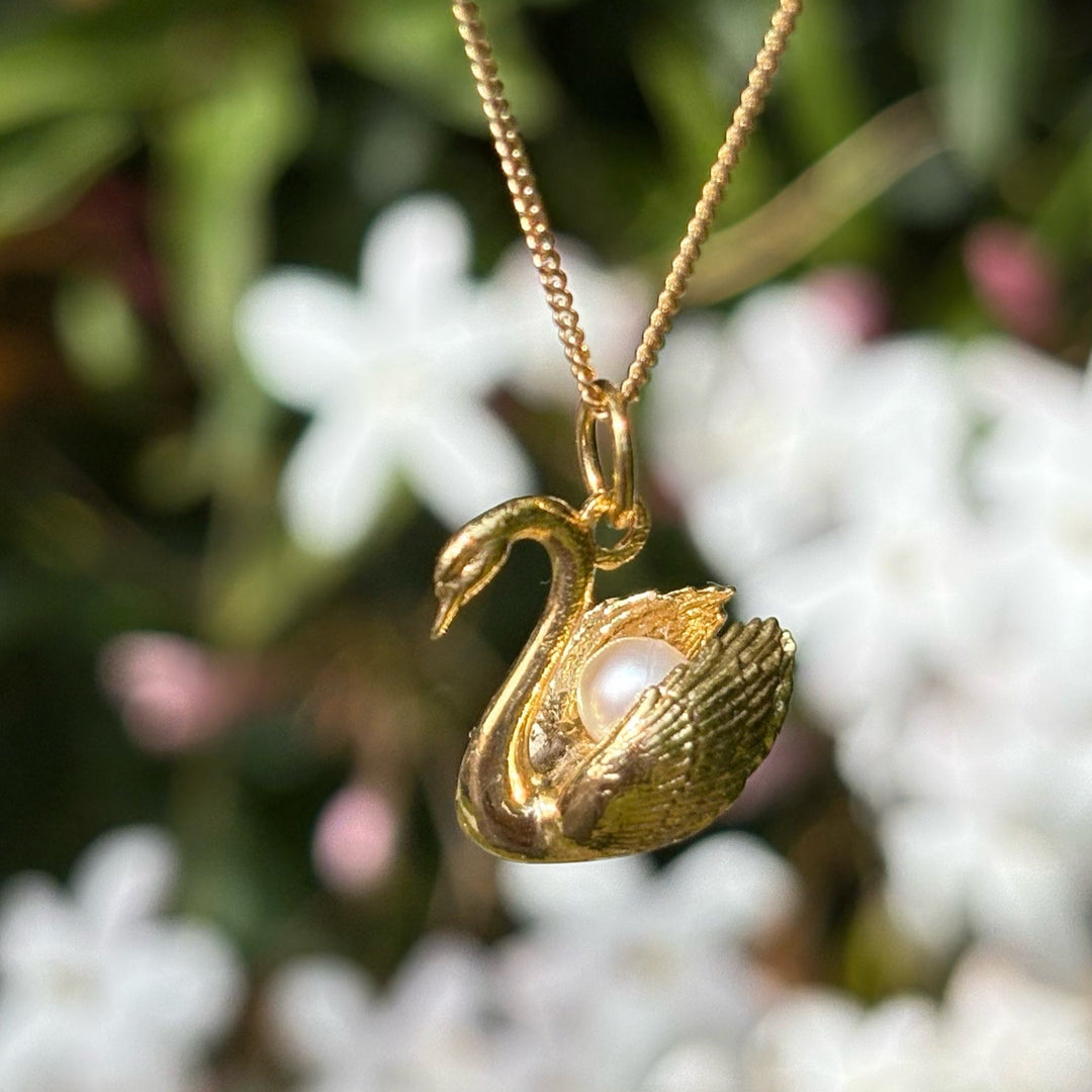 Blackbird Jewellery - A gold vermeil swan necklace with a pearl nestled in its neck, displayed against a blurred background of flowers.