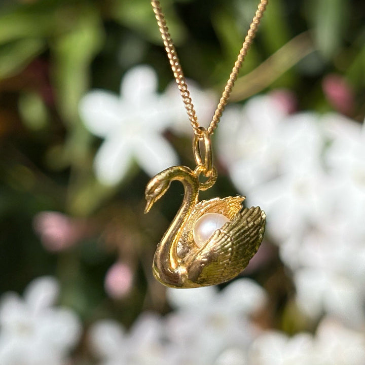 Blackbird Jewellery - A gold vermeil swan necklace with a pearl nestled in its neck, displayed against a blurred background of flowers.