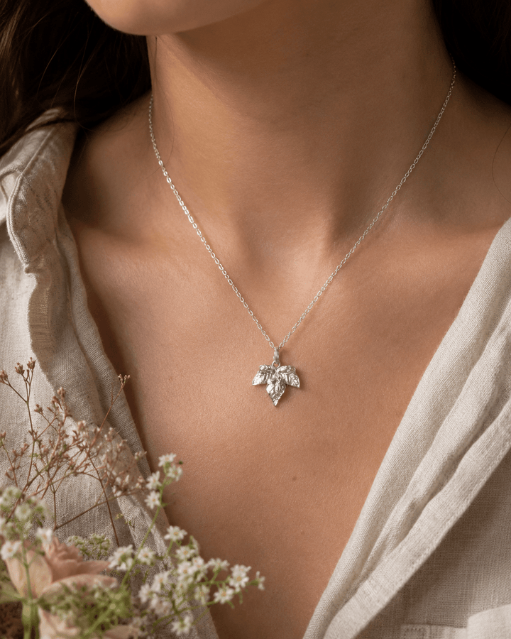 A close-up of a woman wearing a delicate silver chain ivy necklace with a small silver ivy leaf pendant, styled with a light beige linen top and dried flowers in the foreground.