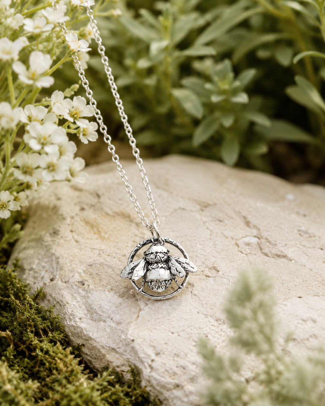 Sterling silver bee necklace styled on natural stone with white flowers and greenery, showing the detailed bee pendant.