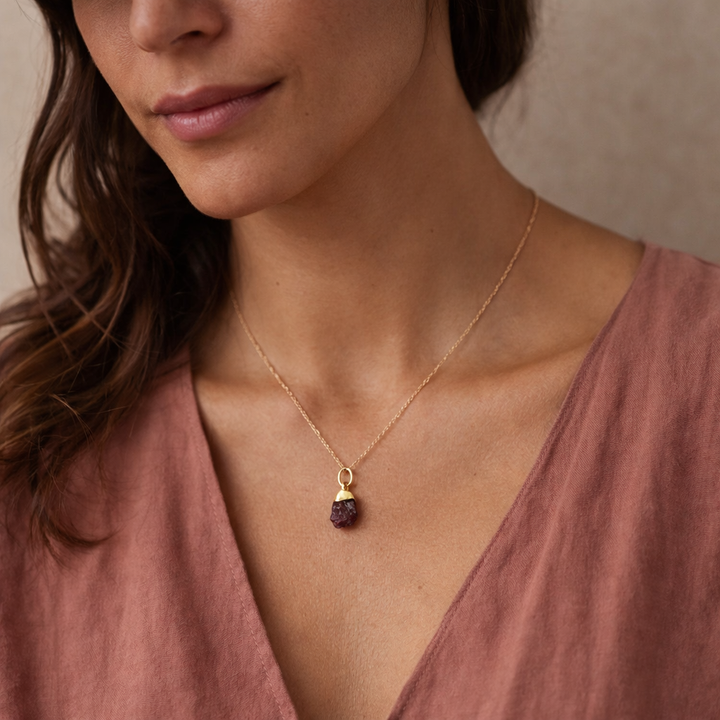 woman wearing a delicate Blackbird Jewellery gold vermeil raw garnet crystal necklace with a deep red-purple rough-cut pendant against a soft warm-toned studio background.