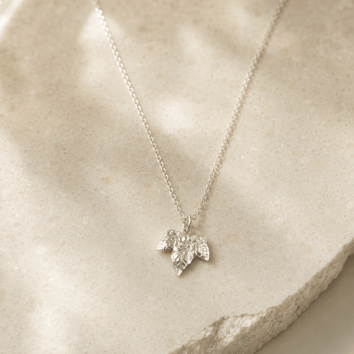A delicate silver chain necklace with a small ivy leaf pendant, photographed on a light beige stone slab with soft shadows.